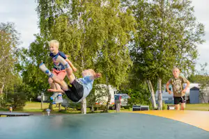 Kinder springen auf einem Trampolin im Campinggelände, im Hintergrund Bäume, Wohnwagen und ein Tisch.