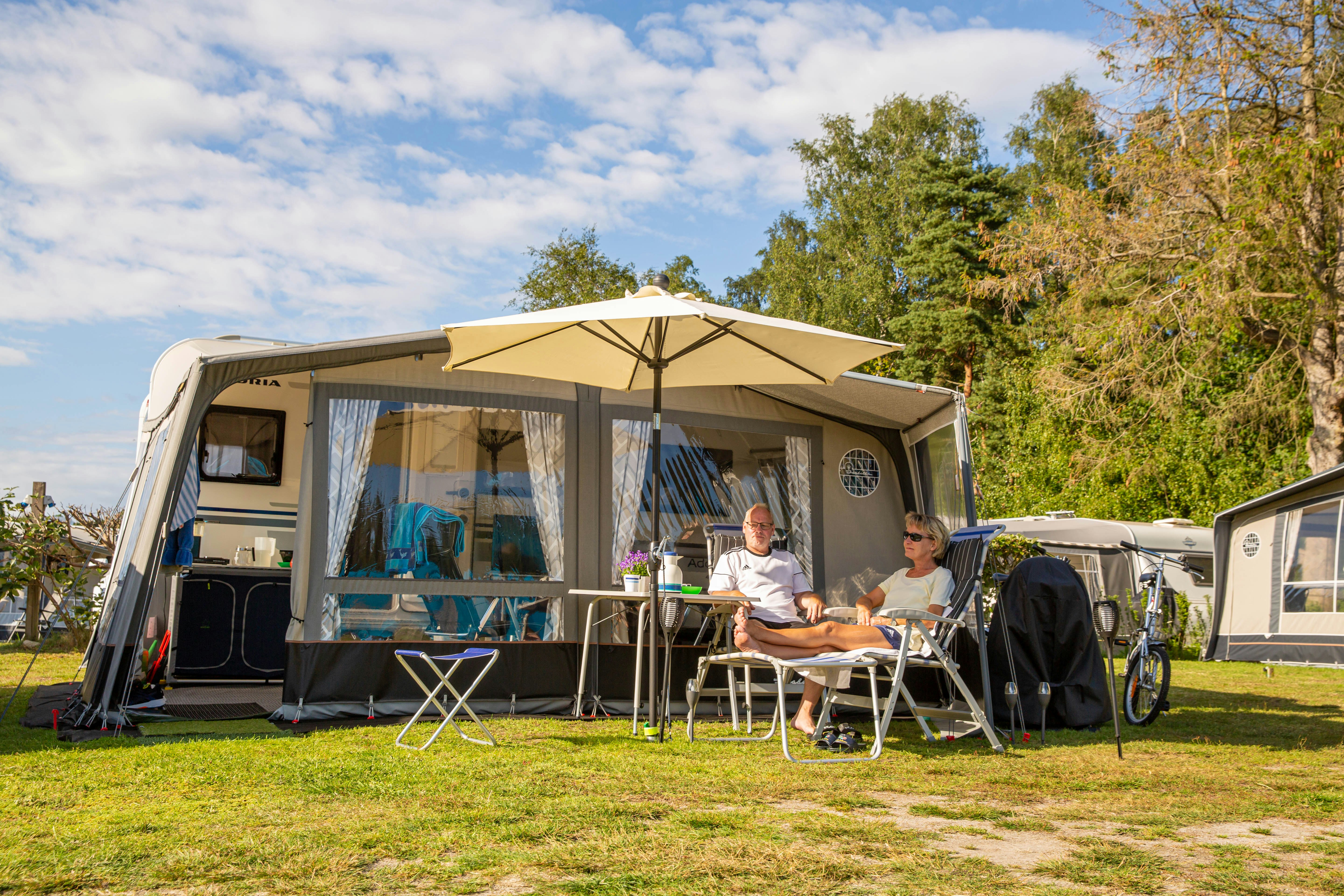 Wohnwagen mit Markise, davor zwei Campingstühle und Tisch auf dem Stellplatz, im Hintergrund weitere Wohnwagen und Bäume.