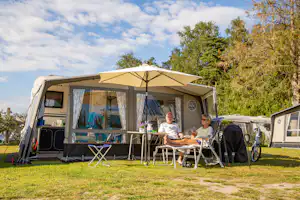 Wohnwagen mit Markise, davor zwei Campingstühle und Tisch auf dem Stellplatz, im Hintergrund weitere Wohnwagen und Bäume.
