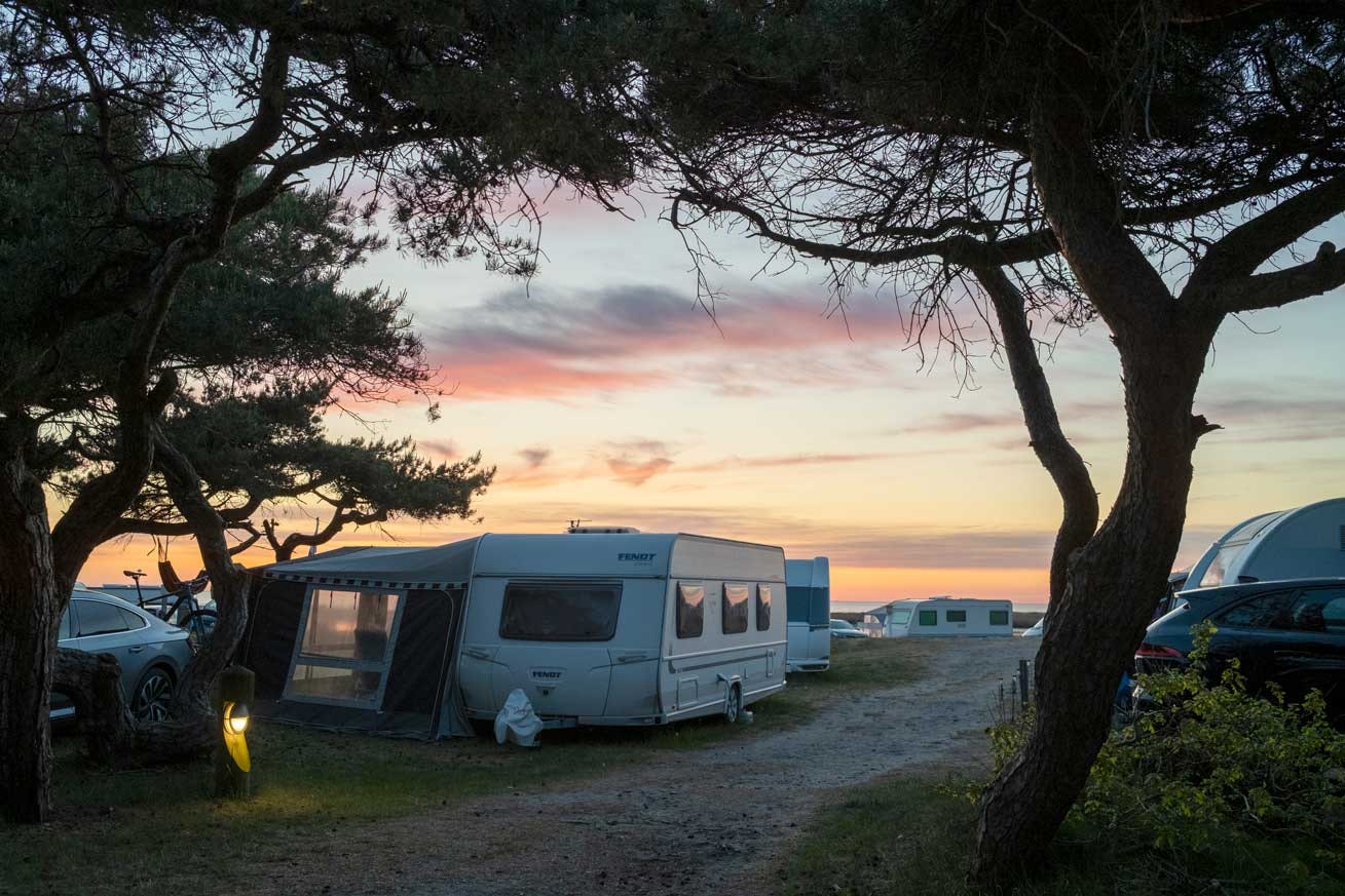 Wohnwagen und Zelte am Campingplatz zwischen Bäumen, im Hintergrund Gebäude und Fahrzeuge bei Sonnenuntergang.