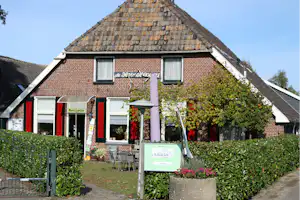 Rezeption-Gebäude aus Ziegeln mit Satteldach, großen Fenstern und Eingangstür, davor Garten mit Hecken und Sitzgelegenheiten.