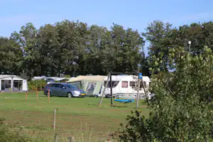 Campingplatz mit Wohnwagen, Zelt und Auto auf dem Stellplatz, umgeben von Rasen und Bäumen.