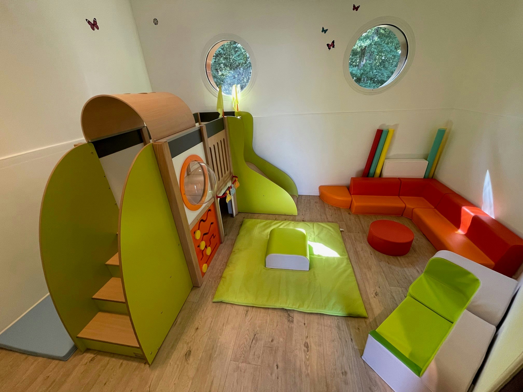 Buntes Kinderzimmer mit Spielzelt, Kissen und Matratze auf dem Boden, daneben ein orangefarbener Sitzbereich.