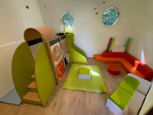 Buntes Kinderzimmer mit Spielzelt, Kissen und Matratze auf dem Boden, daneben ein orangefarbener Sitzbereich.
