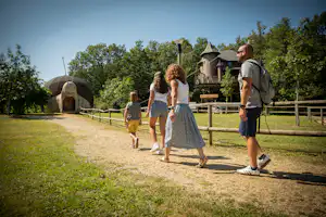 Familie geht auf einem Campingweg, im Hintergrund ein Iglu-Zelt und ein Holzhaus mit Veranda und Geländer.