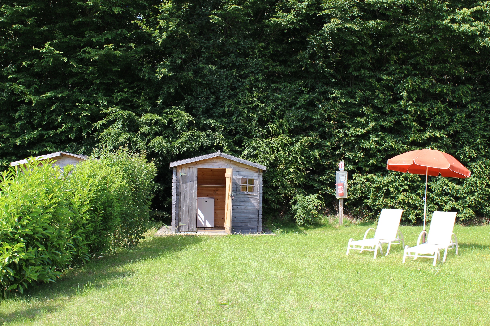 Holz-Saunahäuschen im Garten mit Rasenfläche, zwei Liegestühlen und orangefarbenem Sonnenschirm vor dichtem Grün.