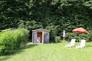 Holz-Saunahäuschen im Garten mit Rasenfläche, zwei Liegestühlen und orangefarbenem Sonnenschirm vor dichtem Grün.