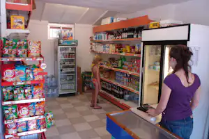 Camping-Shop mit Getränkeregal, Kühlschrank und Snacktheke, daneben steht eine Frau und ein Kind im Innenraum.