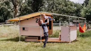 Camping-Unterkunft mit Vorzelt und Holzpodest im Grünen, Person steht davor, im Hintergrund Bäume und Zaun.