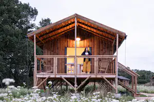 Holz-Campinghaus auf Stelzen mit Balkon, großen Fenstern und warmem Licht, umgeben von Bäumen und Blumen.