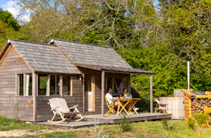 Holz-Cabin mit Veranda und Terrasse, zwei Liegestühle und Tisch, umgeben von Bäumen und Rasen im Campinggarten.