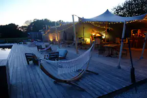 Camping-Terrasse mit Holzplanken, Hängematte, Zelten und Lichterketten, daneben Stühle und Tische am Abend.