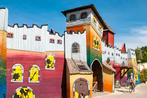Buntes Campinghaus mit gemusterten Türen und Fenstern, Zaun und Gehweg; im Hintergrund steht ein Fahrrad.