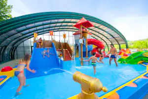 Kinder spielen im überdachten Schwimmbecken mit Wasserrutsche und Spielgeräten, im Hintergrund Bäume und Hügel.