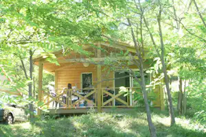 Holzbungalow mit Veranda und Geländer im Grünen, umgeben von Bäumen; daneben steht ein Auto am Weg.