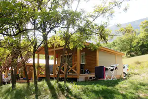 Holz-Chalet mit überdachter Terrasse, Campingstühlen und Markise im Grünen, umgeben von Bäumen und einem geparkten Auto.