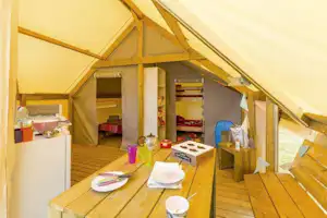 Innenansicht eines Zelts mit Holzterrasse, Tisch mit Geschirr und Küchenutensilien sowie Regalen und Sitzbank.