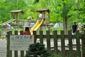 Spielplatz mit gelber Rutsche und Holz-Spielgeräten, Schild im Vordergrund, umgeben von Bäumen und grünem Campinggelände.