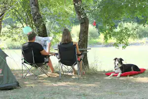 Campingplatz am Fluss: zwei Campingstühle unter Bäumen, Zelt im Hintergrund, Hund liegt auf einer roten Decke.