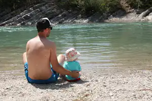Vater und Kinder sitzen am Flussufer im Kies, schauen ins grüne Wasser, im Hintergrund Felsen und Uferböschung.