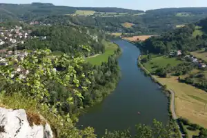 Blick auf einen Fluss im grünen Tal, umgeben von Wiesen, Wäldern und kleinen Häusern am Ufer.