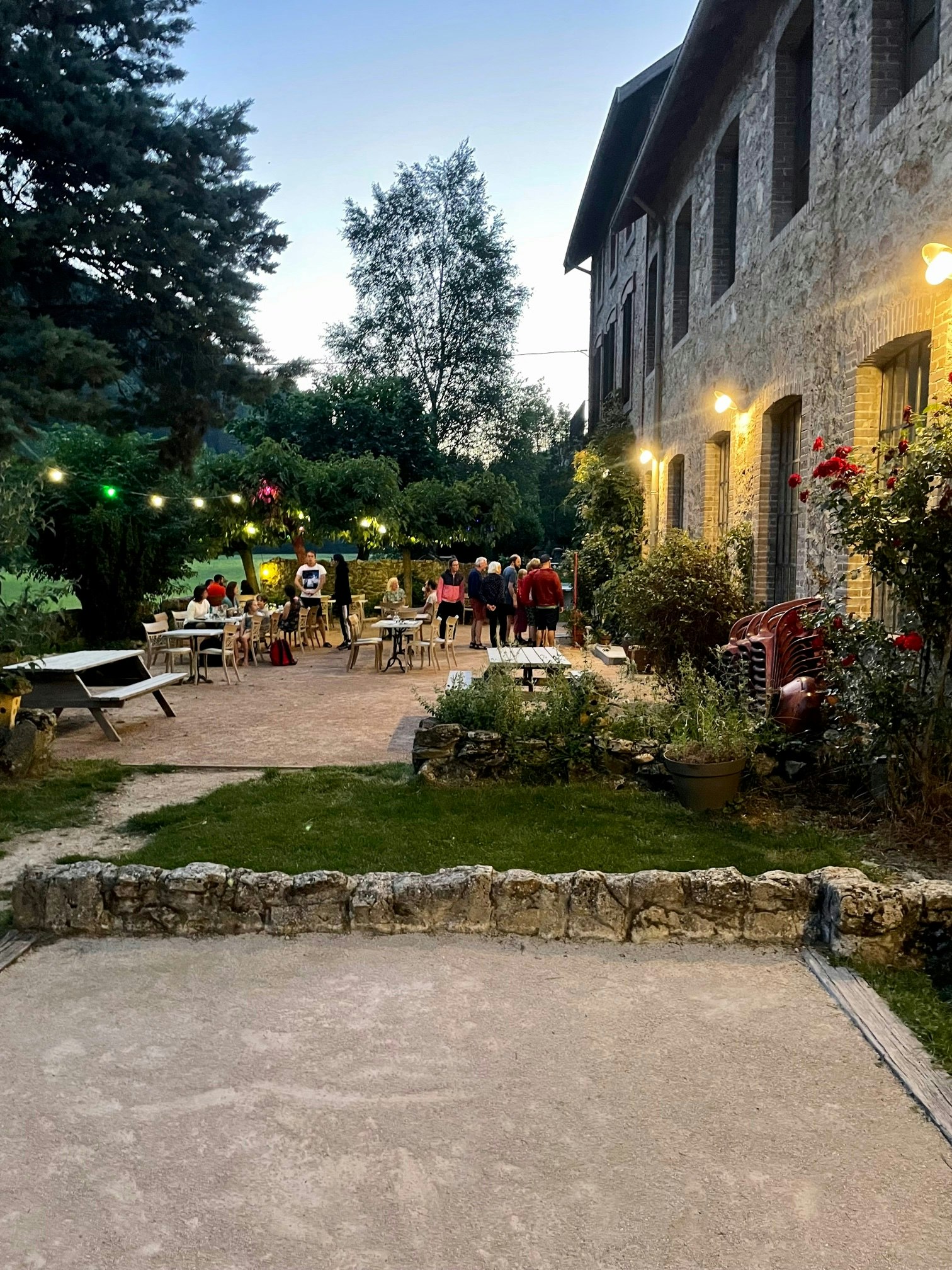Terrasse am Campinghaus mit beleuchteten Tischen, Stühlen und Blumenbeeten, daneben Rasenfläche und Steinmauer im Garten.