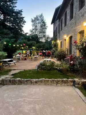 Terrasse am Campinghaus mit beleuchteten Tischen, Stühlen und Blumenbeeten, daneben Rasenfläche und Steinmauer im Garten.