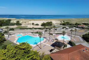 Blick auf einen Campingplatz mit zwei Außenpools, Liegen und Wegen, umgeben von Bäumen und nahe dem Sandstrand am Meer.