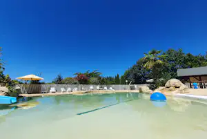 Schwimmbecken mit blauem Planschbecken, Sonnenschirm und weißen Liegestühlen, umgeben von Steinen, Hecken und Bäumen.