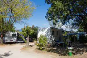 Wohnmobile auf einem Campingplatz zwischen Bäumen, mit Tisch und Stühlen am Stellplatz und einem Weg im Hintergrund.