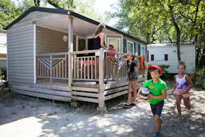 Mobilheim mit Holzveranda und Markise, im Hintergrund weitere Wohnwagen und Bäume im Campingbereich.