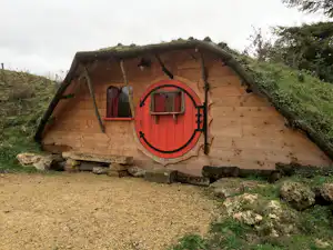 Holzsauna mit roter Tür und rundem Fenster, auf einem gepflasterten Fundament am Hang, umgeben von Gras und Büschen.