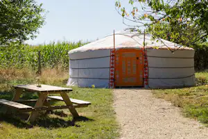 Yurt-Zelt mit Holz-Picknicktisch auf einer Wiese, umgeben von Bäumen und einem Kiesweg.