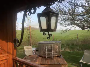 Laterne auf einer Holzterrasse am Campingplatz, daneben ein Feuerkorb und Blick auf Wiese und Bäume.