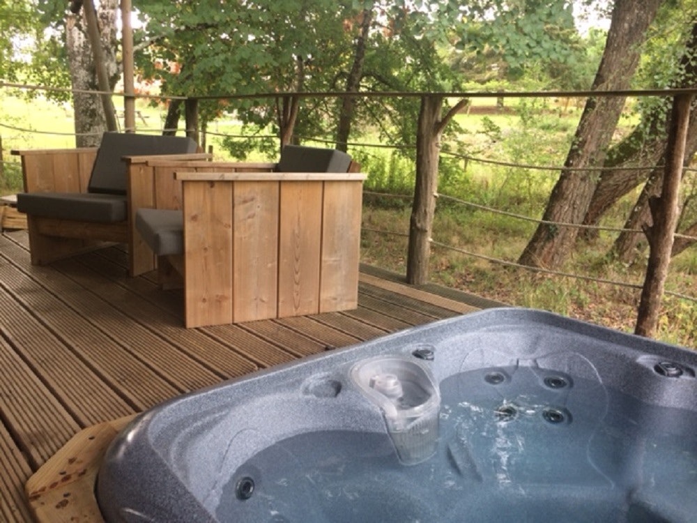 Holzterrasse mit Whirlpool im Vordergrund, daneben Sitzbank und Blick durch den Zaun auf Bäume und Wiese.