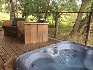 Holzterrasse mit Whirlpool im Vordergrund, daneben Sitzbank und Blick durch den Zaun auf Bäume und Wiese.