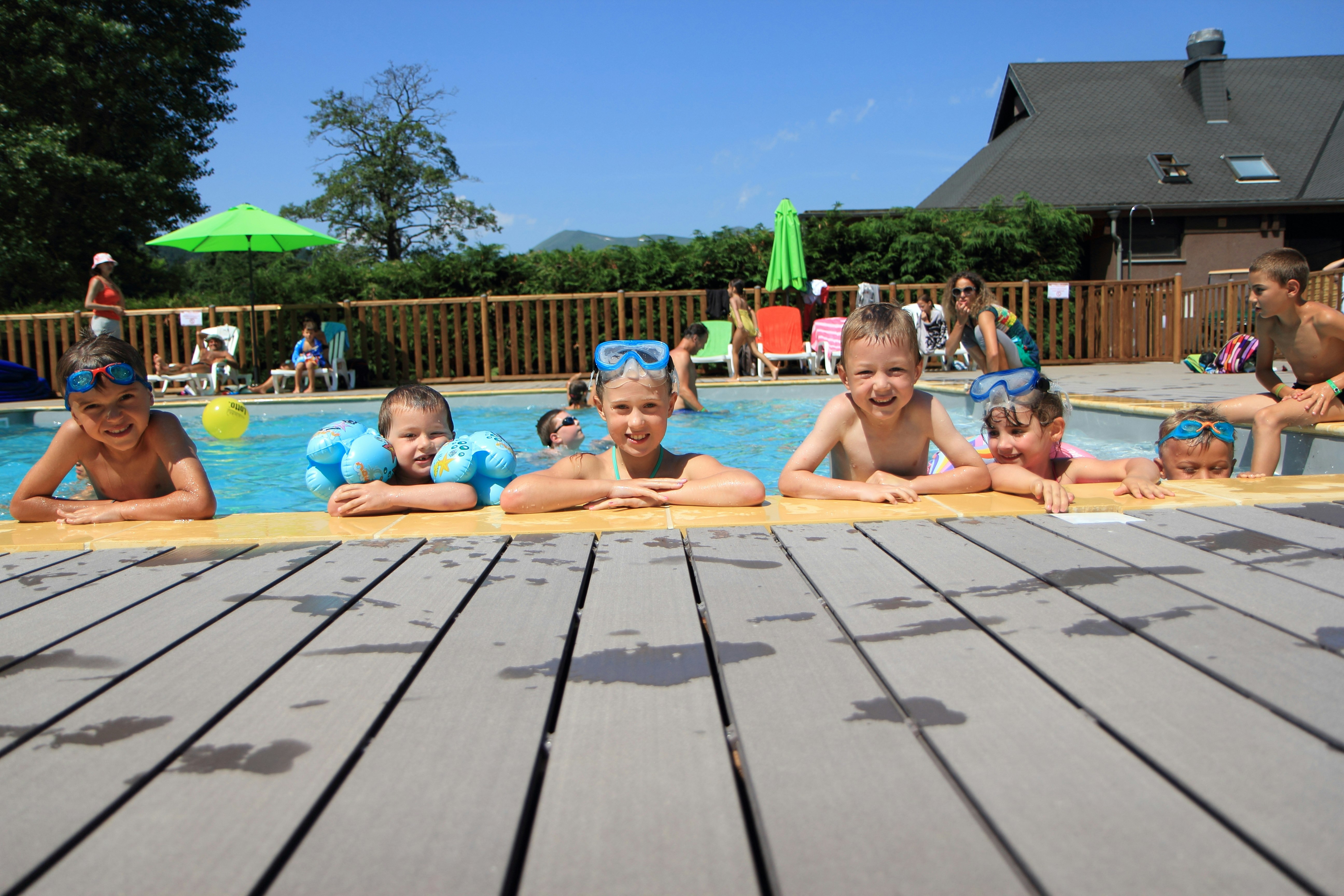 Kinder im Schwimmbecken mit Schwimmreifen vor einem Campinghaus, im Hintergrund Sonnenschirme, Holzterrasse und weitere Familien.