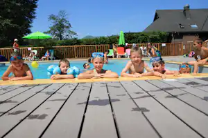 Kinder im Schwimmbecken mit Schwimmreifen vor einem Campinghaus, im Hintergrund Sonnenschirme, Holzterrasse und weitere Familien.