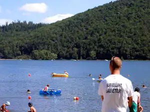 Blick auf einen See mit Schwimmern, einem Schlauchboot und einem gelben Ruderboot vor bewaldeten Hügeln.
