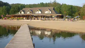 Holzsteg ins Wasser vor dem Sandstrand, dahinter Campinggebäude mit Terrasse und Bäumen im Hintergrund.