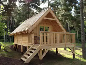 Holzhütte mit Terrasse und Treppe im Wald, umgeben von Bäumen und grünem Rasen.