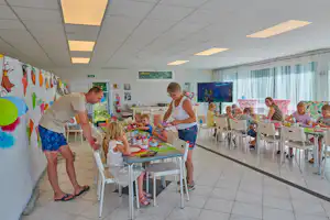 Familie beim gemeinsamen Basteln am Tisch im Gemeinschaftsraum, Kinder mit Farben und Stiften, Erwachsene helfen, große Fenster und Fernseher im Hintergrund.