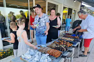 Grillbuffet am Campingplatz: mehrere Personen stehen vor dem Grill und essen an der Terrasse neben dem Gebäude.