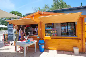 Holz-Verkaufsstand mit Schaufenster und Regalen, davor Tisch mit Getränken und zwei Personen auf dem Campingplatz.