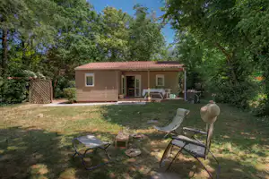Bungalow auf einem Campingplatz mit Terrasse, Gartenmöbeln und Liegestühlen im Grünen, umgeben von Bäumen.