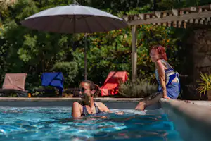 Zwei Personen im Camping-Pool, Sonnenschirm und Liegen im Hintergrund, umgeben von Büschen und einer Steinmauer.