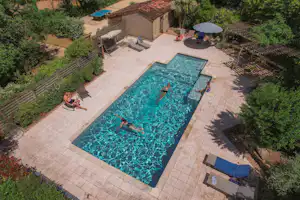 Großer Camping-Pool mit Liegen, Sonnenschirm und gepflasterter Terrasse, umgeben von Bäumen und einem Steinhaus.