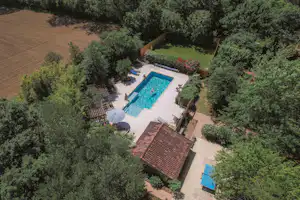 Luftaufnahme eines Ferienhauses mit rotem Dach, Terrasse und blauem Pool im Garten zwischen Bäumen.