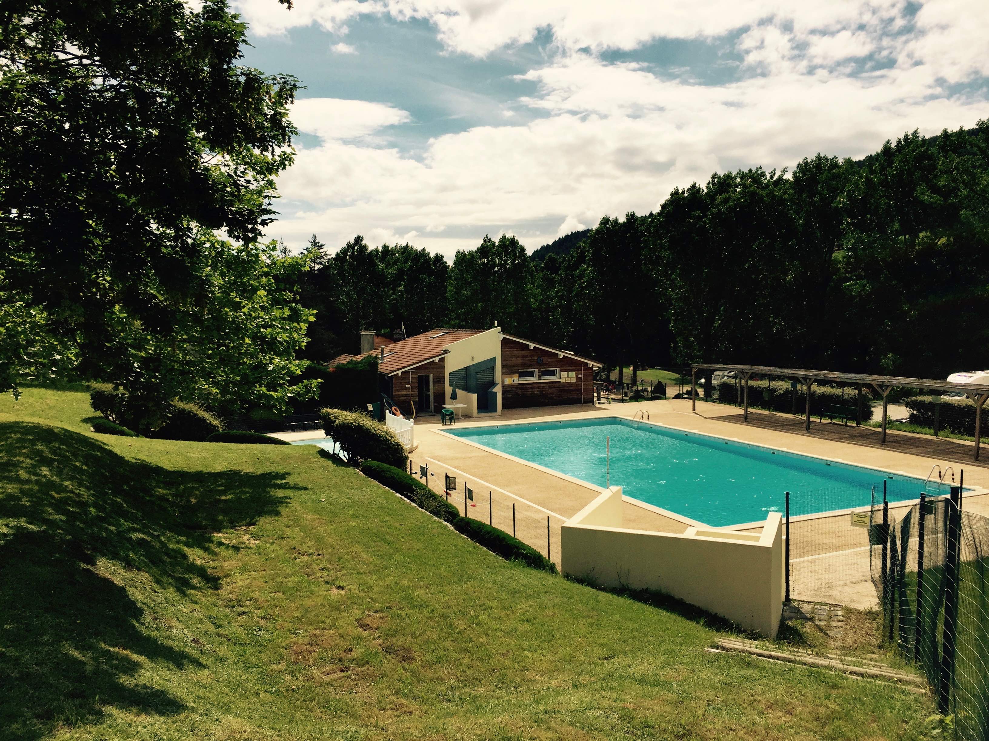 Camping-Pool im Grünen mit Liegewiese, Zaun und einem Gebäude mit Holzveranda am Rand des Geländes.
