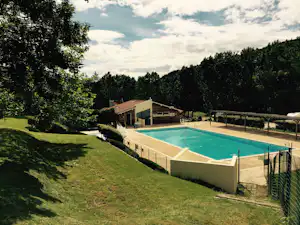 Camping-Pool im Grünen mit Liegewiese, Zaun und einem Gebäude mit Holzveranda am Rand des Geländes.